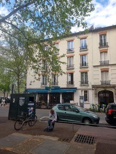 This image captures a street scene in Clichy, France, on what appears to be a bright, clear day with blue skies and scattered white clouds. The primary subject is a multi-story building, light beige in color with brick detailing, featuring multiple windows with dark frames and ornate wrought-iron railings on the upper floors. Some windows have terracotta-colored sills or accents.

At the ground level of the building, there's a commercial storefront with a dark blue awning, which reads "Polette" and "SERVICE CONTINU." To its right is an ornate arched entrance. In front of the building, a street is visible with a light green hatchback car parked along the curb, followed by a black car. Several green shared bicycles are parked further down the street.

On the sidewalk in the foreground, a person wearing a light grey hoodie and dark pants is bending down near a dark-colored bicycle, which is secured to a metal street barrier. The person appears to be interacting with a dark bag or the large dark green collection bin located beside the bicycle. This bin has "LE RELAIS" printed prominently on its sides, along with graphics indicating textile recycling and smaller text that includes "DE FLOUKIER ET TRINQUIER." Another person is visible further down the street on the sidewalk.

A large, leafy green tree partially obstructs the view of the building on the left, its branches and foliage framing the top and left portions of the scene against the sky. A street drain cover is visible on the sidewalk in the lower right corner. The overall setting is an urban environment, likely a residential and commercial street.