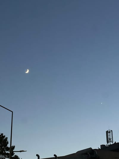 The image depicts a twilight sky over Năvodari, Romania. A crescent moon is visible in the upper left quadrant of the sky. To the right of the moon, a bright star or planet is also visible. In the foreground, silhouetted against the sky, are the rooftops of buildings. A large antenna tower is prominent on the right side of the image, situated on a rooftop. On the left, a streetlight and the dark foliage of a tree are partially visible. The overall environment is urban, captured during the transition from day to night.