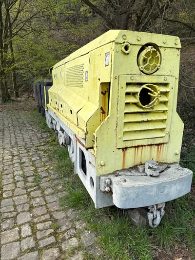 The image displays a static, weathered, bright yellow industrial mining locomotive positioned outdoors on a grassy verge adjacent to a cobblestone path. The front of the locomotive features a robust, light gray bumper. Above the bumper, the yellow body includes a horizontal grated section, a circular fixture with a spoked interior, and a circular opening below the grate, featuring bent metallic components within. Prominent rust streaks are visible below the grated section and along various seams on the locomotive's body. The chassis and wheels are predominantly light gray. Partially visible behind the main locomotive body is a dark-colored wagon, likely a small coal cart. To the left, a cobblestone path runs parallel, with green grass and several yellow dandelions growing between the path and the train. The background comprises a forested hillside with trees exhibiting light green, nascent spring foliage. No individuals are present in the frame, and no active operations are depicted. This scene is located at the LWL-Industriemuseum Zeche Nachtigall in Witten, Germany.