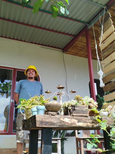 A man wearing a yellow bucket hat and a blue t-shirt stands in front of a white building with red window frames. He is smiling and looking at the camera. To his right, there is a wooden table with various potted plants, including succulents, a decorative candelabra, and a macrame plant holder hanging from the roof. The building appears to be a residence or shop in Trường An, Vietnam, with a corrugated metal roof supported by red beams. The overall setting suggests an outdoor area, possibly a porch or garden, decorated with plants and rustic furniture.