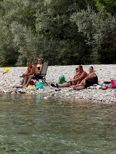 The image shows a group of people relaxing on a pebbly beach next to a calm body of water, likely the river Vrbas near Spiljani, Bosnia and Herzegovina. The sun is bright, suggesting a warm, clear day. The group appears to be enjoying a leisurely day by the water.
In the foreground, two men are seated on the beach. One man, facing the camera, is wearing black shorts and has his arm around another man who is also in shorts. They seem to be engaged in conversation or simply enjoying each other's company. Behind them, a woman in a black swimsuit with purple accents is seated in a beach chair, looking towards the water. She is wearing sunglasses.
Further back on the beach, a group of three men are sitting together, also on the pebbles. They are all wearing swim trunks. One man is leaning against another, and they all appear relaxed.
Scattered on the beach are various objects, including a green bag, a red towel, and several plastic bags filled with what appear to be trash or belongings. There is also a folded beach chair and other folded fabric items. The water is clear, with sunlight reflecting off its surface, creating a shimmering effect. The background is dominated by lush green trees, indicating a natural, possibly forested, environment surrounding the beach. The overall mood is one of relaxation and enjoyment of a summer day outdoors. No visible text is present in the image.