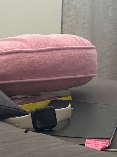 The image shows a close-up of a person's lap with a pink garment draped over it. To the right, a black smartwatch with a white band is visible, attached to a grey jacket sleeve. Behind the smartwatch, a black notebook or folder is propped up, with a piece of pink sticky note attached to its edge. A sliver of a yellow object is visible between the pink garment and the notebook. The background is out of focus, appearing to be a textured grey surface. The lighting suggests it could be daytime indoors. No weather or specific location cues are discernible beyond the provided context of Hausach, Germany. No discernible activity is taking place.