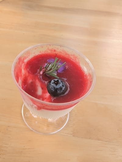 A close-up, eye-level shot shows a single serving of panna cotta in a clear plastic cup, placed on a light-colored wooden surface. The dessert has two distinct layers: a white, creamy base topped with a vibrant red berry sauce. A single, dark blue blueberry sits in the center of the sauce, and a sprig of rosemary and a small purple edible flower are artfully placed on top of the blueberry and sauce. The wooden surface has a visible grain pattern and appears to be a tabletop in a cafe or restaurant in Tambon Mae Raem, Thailand. The lighting is natural and bright, highlighting the textures and colors of the dessert.