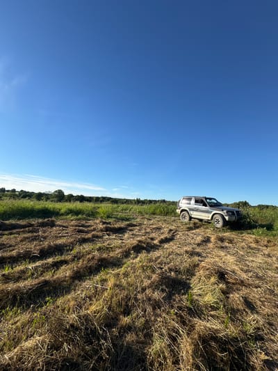 A silver, two-door SUV is parked in a field of dry, cut grass under a clear blue sky. The SUV is facing right, with its front slightly angled towards the viewer. The field has visible tire tracks from the vehicle. In the background, there is a dense line of green trees and vegetation. The scene appears to be in a rural area.