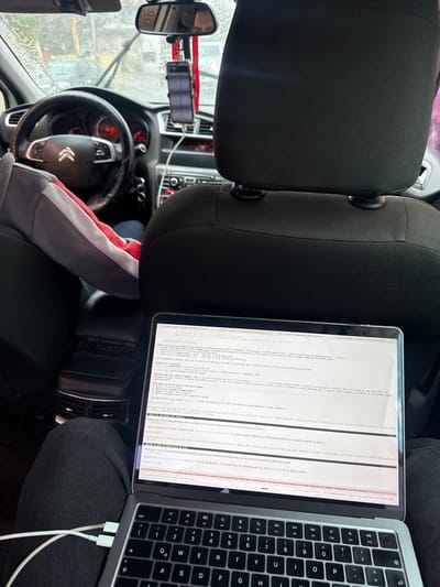 The interior of a Citroen car is visible, featuring a black seat with a headrest. A laptop rests on the passenger seat displaying code or documentation, and a smartphone is mounted on the windshield, potentially used as a GPS device. The steering wheel with the Citroen logo is visible, and the overall environment suggests the car is parked, possibly during a pause in a journey, in Quilmes Oeste, Argentina.