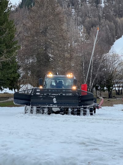 A black snow groomer, identified as a "LEITWOLF," is driving on a snow-covered road in Val-d'Isère, France. The groomer is facing the camera and has a large snow blade attached to its front. The groomer is driving on a snow-covered road. The road is surrounded by snow-covered trees and shrubs. A red object is visible on the right side of the groomer. The road is slightly sloped and the groomer is moving forward. The driver can be seen through the front window. A second, smaller, orange object is visible at the top of the image. The image is taken from a low angle, looking up at the groomer.