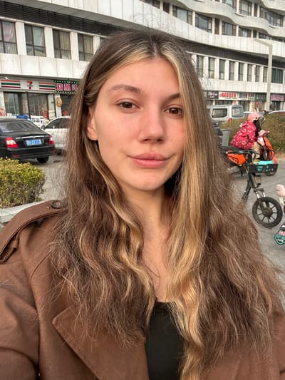 A female with light skin, brown eyes, and long, wavy brown hair featuring blonde highlights poses for a photograph. She wears a brown, collared coat with shoulder epaulets over a black garment, exhibiting a neutral expression with a slight smile. The scene is an urban streetscape in Huangdao District, Qingdao, Shandong Province, China.
In the background, a 7-Eleven convenience store, identifiable by its green, red, and white logo and bilingual signage (English and Chinese), is visible. Parked on the street in front of the store is a black sedan, specifically a Hyundai, displaying the license plate "鲁U 22713," indicating its registration in Shandong province, with 'U' designating Huangdao District. A white car is partially visible behind it. Green bushes in planters line the sidewalk.
To the right, an electric bicycle equipped with an orange cargo rack is parked on the sidewalk. A small child, dressed in a red patterned jacket and a pink helmet, is seated in a rear-mounted child seat on the bicycle. Multi-story buildings with a light-colored facade and distinctive wavy architectural elements on upper floors form the backdrop. The lighting indicates daytime conditions.