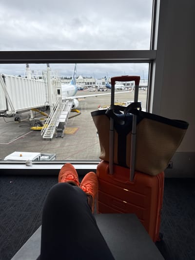 A person's legs, wearing bright orange sneakers and black pants, are visible in the foreground, positioned to the right of an open window. The window offers a view of an airport tarmac. An orange suitcase and a woven straw tote bag with dark straps rest on the floor next to the window.
Through the window, a jet bridge is connected to an airplane. The plane's tail, which has a blue and green logo, is visible. Various airport vehicles and equipment are also present on the tarmac. The sky appears overcast, suggesting a cloudy day. The scene is set at an airport gate, likely in SeaTac, USA, as indicated by the location context, and the perspective suggests the person is waiting to board a flight. The overall mood is one of anticipation for travel. No visible text is present in the image.
