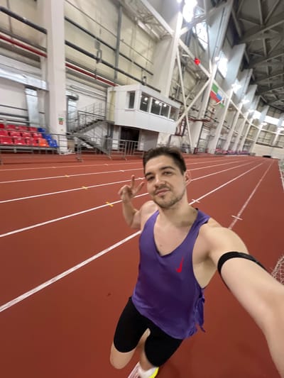 An adult male is captured in a selfie within an indoor athletic stadium, located in Tashevka, Russia. The man, with light skin and dark hair, is wearing a purple Nike tank top, black athletic shorts, and athletic shoes with white soles and neon yellow/green accents. A black armband is visible on his left upper arm. He looks directly at the camera with a slight smile, his right hand raised to display a peace sign.

The background features a red-surfaced athletic track with multiple white lanes and yellow markings, extending into the distance. To the left, rows of red and blue plastic stadium seats are visible below a white, elevated control booth with large windows, which is accessible via a metal staircase. The facility's high ceiling is supported by white metal trusses and pillars, with silver ventilation ducts also visible. Two flags hang from the ceiling structure on the right side of the image: the national flag of Russia and the flag of Tatarstan. A white metal barrier separates the track from the seating area and control booth access. No other individuals are present in the visible area.