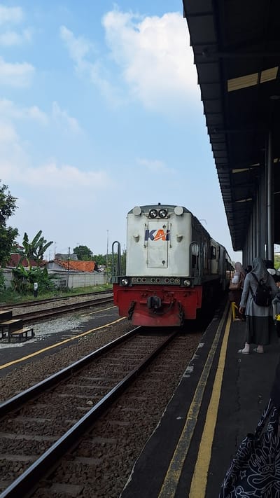 The image depicts a train at a station, possibly in Karawang, Indonesia. A white and red locomotive with the "KAI" logo is positioned on the tracks, facing the viewer. The train is at a platform where several individuals, dressed in modest attire, are waiting. One person, wearing a gray hijab and backpack, stands out. The station features a covered walkway with visible support pillars and a dark roof. The sky is clear with scattered clouds, and in the background, a residential area with houses and vegetation is visible. The ground is covered with gravel and ballast, typical of railway infrastructure. The scene appears to be in the daytime under natural light.