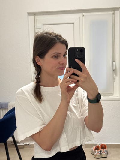 A young woman is taking a selfie in a bathroom. She is holding a black smartphone with multiple camera lenses in her right hand, her face partially obscured by the phone. She has brown hair partially tied back with a scrunchie, blue eyes, and fair skin. She is wearing a white t-shirt and a smartwatch with a green band. Behind her is a bathroom mirror and white wall. On the counter above, the mirror reflects a pair of orange and white sneakers. The location appears to be indoors in Kőröshegy, Hungary.