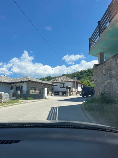 The image shows a view from inside a vehicle, possibly a car, looking out onto a street in the village of Leshten, Bulgaria. The sky is blue with scattered white clouds. Traditional stone houses line the left side of the street, constructed with white walls and stone roofs. A black SUV is parked further up the street. There are green trees and vegetation in the background. Power lines are strung across the sky. Part of the vehicle's dashboard is visible at the bottom of the image, and part of an overhanging structure appears at the top left. The road is paved and appears to be mostly empty.