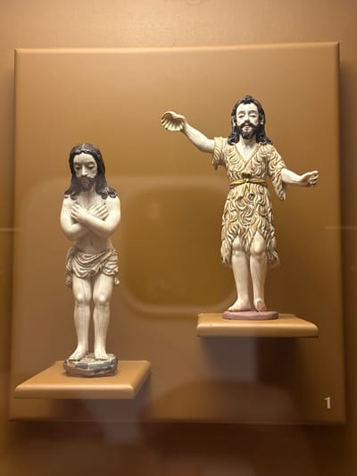 The image displays two intricately carved religious statues, likely from a museum or exhibition in Tepotzotlán, Mexico. Both figures are male with long, dark hair and beards, and are presented against a plain, light brown background, possibly behind a glass barrier given the faint reflections.
The upper statue depicts a figure standing upright, with his right arm raised and his left hand holding a shell. He wears an off-white, textured garment, possibly resembling animal skin or fur, with golden accents around the waist and collar. This figure's iconography, particularly the shell, suggests a depiction of Saint John the Baptist. His feet are placed on a reddish-purple element set on a small, light-brown rectangular base.
The lower statue portrays a figure lying in repose or deceased, with his eyes closed and hands crossed over his chest. He is partially draped in an off-white garment with some gold detailing. Visible on his bare chest and legs are delicate carved lines, possibly anatomical or representing wounds. This figure could represent the Dead Christ or a recumbent saint. His feet rest on a blue-grey element atop a similar small, light-brown rectangular base.
The statues exhibit fine detail in their carving, particularly in the hair, beards, and garments. A white numeral "1" is visible in the top right corner of the background, possibly an exhibit number. The overall display is simple, focusing attention on the figures themselves.