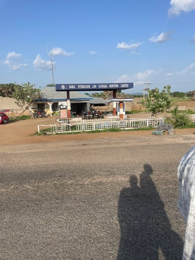 This media file captures a roadside view in Wai A, Nigeria, under a clear, bright blue sky with scattered white clouds, suggesting a sunny day. The scene is dominated by a gas station with a prominent blue sign reading "HARA PETROLEUM AND GENERAL SERVICES LIMITED." The station appears to be operational, with motorcycles parked around the building, indicating a degree of local activity.
Several people are visible, some near the gas station, suggesting they are customers or employees. The presence of a red car parked at the bottom left corner further suggests everyday use of the location. In the foreground, the texture of an asphalt road is visible, with the shadow of what appears to be a person casting a distinct silhouette on the surface, hinting at the time of day being midday or early afternoon.
Further details include a white metal fence surrounding parts of the gas station, a tall antenna structure with multiple components, and lush green trees and vegetation, which are characteristic of the Nigerian landscape. The overall impression is that of a typical day at a rural or semi-urban gas station, serving the local community.
