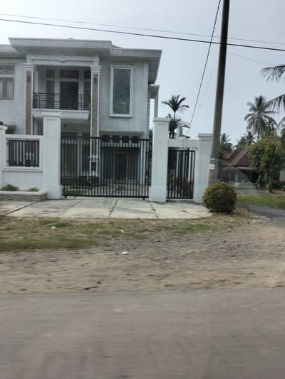 The image depicts a modern, two-story house with a white exterior, black metal gates, and a long driveway. The house features a balcony on the second floor with a black metal railing, a large window with a diamond pattern on the right side of the facade, and several other windows and doors. A black metal fence runs along the front of the property. Palm trees are visible in the background, along with a separate, older-style house with a reddish-brown roof. The scene is set on a clear day, with a cloudy sky. The foreground shows a rough, gravelly road. The location is identified as Tingkeummanyang, Indonesia. There are no people or animals visible in the image, nor are any actions or interactions taking place. The overall impression is one of a suburban or semi-rural residential area.