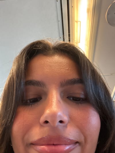 The image presents a close-up, low-angle view of a single individual's face, positioned within what appears to be the interior of a public transit vehicle in Linden, United States. The subject has medium-dark brown hair, dark eyes, and a medium skin tone. Facial features including the forehead, eyebrows, eyes, nose, and lips are discernible; the lips exhibit a glossy texture. The individual's gaze is directed downwards. The background, appearing out of focus, reveals the utilitarian interior architecture of the vehicle, characterized by light-colored paneling with visible seams. A vertically oriented, illuminated fluorescent light fixture is prominent in the upper right quadrant of the frame, with a partially visible circular fixture further to the right. No other individuals or direct interactions are visible.