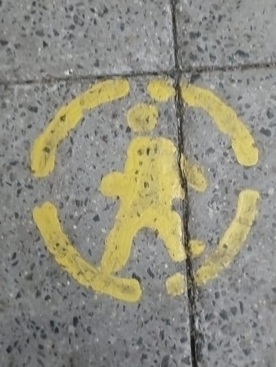 The image shows a yellow painted symbol on a textured, grey concrete surface. The symbol depicts a stylized human figure walking within a broken circle. The figure appears to have a head, torso, and legs, with one leg forward and the other back, indicating movement. The circle is formed by two curved lines, each with a gap in the middle, suggesting a path or a designated area. The concrete itself is composed of small, dark grey and light grey aggregates embedded in a lighter grey matrix. There are visible seams or grout lines running vertically and horizontally through the concrete, indicating it is laid in sections. The painting is somewhat faded and worn, with parts of the yellow paint chipped or smudged, suggesting it has been exposed to the elements or foot traffic. The lighting is even, with no strong shadows, suggesting it might be an overcast day or the image was taken indoors. The setting appears to be a pedestrian area, possibly a sidewalk or a path, given the painted symbol and the concrete material. No text is visible in the image.