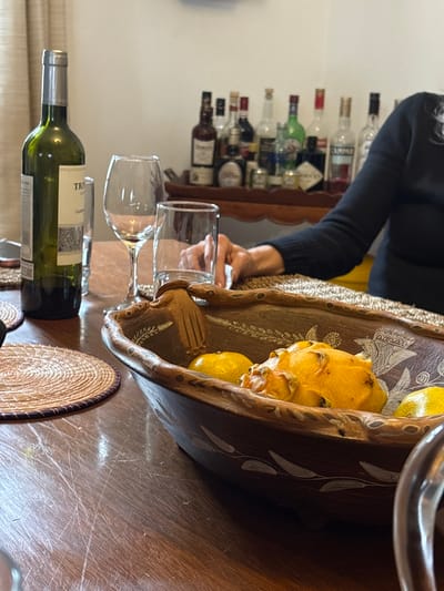 The image shows a still life on a wooden table, likely indoors. There is a bottle of white wine lying horizontally, two wine glasses, a large, shallow wooden bowl with fruit (possibly lemons and a dragon fruit), and a circular woven mat. A person wearing a dark long-sleeved shirt is partially visible in the background, near what appears to be a liquor shelf. There is no verifiable information to conclude that the image was taken in the city of Aucallama, Peru.