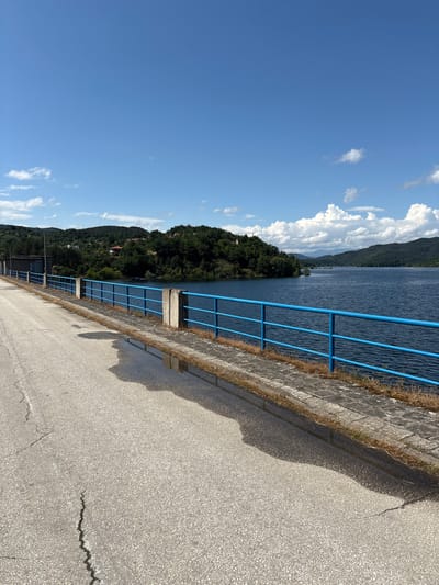 This image captures a bright, sunny landscape overlooking a large body of water, likely a reservoir near Muhovo, Bulgaria. The scene is static, with no discernible activity or people.

In the foreground, a wide, light-colored paved surface, appearing to be concrete or asphalt, stretches across the frame. It shows signs of wear with several visible cracks, including a prominent zigzag crack. Dark shadows, likely cast by the railing and an unseen object (possibly the photographer), extend across the paved area. To the left of the paved surface (when viewed upright), there is a low wall constructed from rectangular grey blocks, with dry grass and weeds growing along its upper edge. A bright blue metal railing, consisting of vertical bars and horizontal supports, runs along the length of this wall, separating the paved area from the water. It is anchored by sturdy posts, some appearing to be wood or concrete.

Beyond the railing and wall lies a vast expanse of deep blue water, exhibiting gentle ripples on its surface. In the midground and background, dense green forested hills and mountains rise from the water's edge, stretching into the distance. Some small structures with reddish roofs are sparsely visible among the trees on the lower slopes of the hills. The sky above is a clear, vibrant blue, dotted with scattered, fluffy white cumulus clouds, particularly clustered towards the upper left.

The time of day appears to be midday or early afternoon, indicated by the strong sunlight and well-defined shadows. The weather is clear and sunny. There is no visible text in the image. The setting suggests an elevated viewpoint, possibly from a dam or embankment, overlooking a scenic natural landscape.