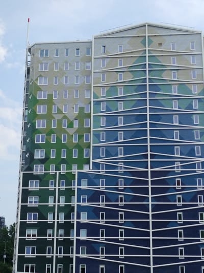 This is a medium shot of a tall, modern building with a colorful, geometric facade. The building is predominantly white with sections of green and blue diamond-shaped patterns that create a gradient effect. White horizontal lines crisscross the facade, creating a sense of depth and division between floors. Numerous rectangular windows, mostly appearing dark or reflecting the sky, are evenly spaced across the building's surface. A thin, white pole with a red tip extends vertically from the top left section of the building.
The scene appears to be outdoors, under a lightly cloudy sky. The lighting suggests it is daytime, possibly late morning or afternoon. The architectural style points to a contemporary urban setting, and the provided location context places it in Zaandam, Netherlands. There are no people or specific activities immediately apparent; the focus is solely on the building's design. No visible text is present in the image. The overall impression is one of modern, vibrant architecture set against a natural sky.