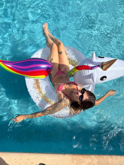 A woman with extensive tattoos on her arms and legs lies on her back on an inflatable unicorn float in a swimming pool. She is wearing a red and white checkered bikini and black sunglasses. The pool is filled with clear blue water. The unicorn float is white with a golden horn and black eyes. A rainbow-colored inflatable is attached to the front of the unicorn. The scene is lit by bright sunlight, casting shadows on the water. The location is the city of Sant Vicenç de Montalt, Spain.