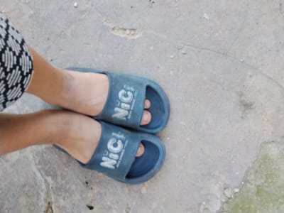 A high-angle, close-up shot shows a pair of blue slide sandals on a cracked concrete surface. The left sandal is on a person's left foot, partially visible, wearing a black and white patterned sock or shoe cuff. The right sandal is on the person's right foot, with the toes visible. Both sandals have the white text "NiC!" printed on them. The concrete has patches of green moss and is generally worn and uneven, suggesting an outdoor environment, possibly in the city of Cîrpești, Moldova.