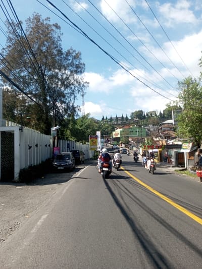 A daytime street scene in Ban Phonhxavanh, Laos, depicts an asphalt road ascending slightly into the distance under a blue sky with scattered white clouds. Multiple dark utility lines crisscross overhead. A solid double yellow line marks the center-right of the road, which is occupied by at least six motorcycles moving away from the viewer. The lead motorcyclist wears a red jacket and a white helmet, carrying a passenger also in a white helmet. On the left side of the road, a long, light-colored concrete wall parallels the street, behind which a large tree with sparse foliage and a communication tower are visible. Two dark-colored sedans are parked parallel to this wall. On the right, various commercial and residential structures line the street, with additional buildings featuring distinct pointed roof structures nestled among dense trees on a rising hillside in the background. Long, linear shadows from overhead lines and objects are cast across the road. A partially visible individual stands on the far right roadside, holding a red bag or basket.