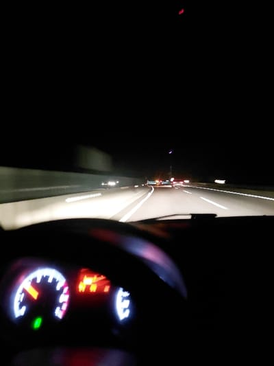 A nighttime view from inside a vehicle shows a dark highway ahead. The road markings are visible, illuminated by headlights. Other vehicles can be seen in the distance with illuminated lights. The interior of the car is visible in the foreground including the steering wheel and dashboard. The dashboard shows illuminated indicators, displaying the speed and other engine details.