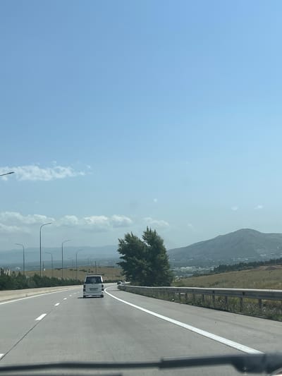 This image shows a view from a vehicle traveling on a highway. The highway has multiple lanes and is bordered by a guardrail on the right. In the foreground, the road surface is clearly visible with white lane markings. A white van is traveling in the same direction as the viewer, in the lane to the left. In the distance, there are several streetlights along the side of the road, and a large green tree is visible on the right. Beyond the highway, there are rolling hills and mountains, with some buildings and structures visible in the valley below. The sky is clear blue with a few scattered clouds. This scene is consistent with the highway leading to Tedotsminda, Georgia, given the landscape and infrastructure.