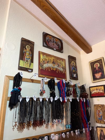 The image depicts a shop interior in Moni Archaggelou, Greece, located at 40.597 latitude and 24.702 longitude. The shop is adorned with traditional religious icons on the walls.  The wall is painted white with a brown wooden beam running diagonally across the top of the wall. The icons are displayed in wooden frames, depicting religious figures and scenes. Below the icons, a wooden rack displays numerous strings of prayer beads, known as komboloi, with price tags attached. The komboloi are arranged in rows and vary in color, style, and size.  The price tags range from 3.00 to 5.50 euros.