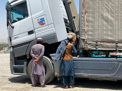The image shows a white semi-trailer truck parked on a dirt road. The truck is a Mercedes-Benz 1841 model, with the words "Mercedes-Benz" visible on the side. The trailer has a "KOEL" logo and a large, gray tarp. The truck is parked in the city of Panjwai, Afghanistan, at the coordinates 31.614, 65.588. Two men are standing beside the truck. Both men are wearing traditional Afghan clothing: long shirts and pants. One man is wearing a white cap and the other is wearing a brown waistcoat.  They appear to be talking to each other. The sky is bright and clear, with no clouds visible. There is a small, green bush in the background on the left side of the image.