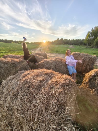 The image captures a serene rural scene at sunset in Стеклянный, Russia, featuring two women amidst several large, round hay bales. The sun, positioned low on the horizon to the left, casts warm, golden light and prominent lens flares across the field, signaling the golden hour. The sky above is clear blue with delicate wispy clouds.

On the left, a woman with light-colored hair, dressed in a dark jacket and pants, sits casually on top of a hay bale. She raises her left arm high, holding a blue-cased smartphone, possibly taking a photo of the sky or a selfie. Her colorful striped socks add a touch of vibrancy to her dark attire. To the right, another woman with light brown hair stands between two hay bales. She wears a pink bomber jacket over a dark top and light blue denim shorts, holding a dark-cased smartphone in front of her, smiling gently as if taking a picture or video.

The hay bales, made of dried straw, fill the foreground and midground, their textures highlighted by the low sunlight. Behind the women and the bales, a vast green field stretches towards a dense line of trees, indicating a forest on the horizon. The setting evokes a peaceful, relaxed atmosphere, suggesting an enjoyable evening spent outdoors. There is no visible text in the image.
