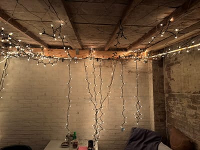 The image depicts an indoor scene, likely a basement or attic space, characterized by exposed wooden beams and a brick wall. The space is adorned with numerous strings of white fairy lights, which are draped across the ceiling and hang down the wall, creating a warm, ambient glow. On a table in the foreground, several objects are visible, including mugs, glasses, a bottle, and a small dish. A cushion or pillow is also present in the lower right corner of the image. The overall impression is one of a cozy, decorated space. There is no discernible location in the city of Boxtel, Netherlands.