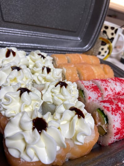 This media file features a close-up, top-down view of a selection of sushi rolls arranged in a black plastic container. The sushi is presented in two distinct rows. The row in the foreground is adorned with dollops of white cream, possibly mayonnaise or a cream cheese frosting, piped in a decorative swirl pattern. Each dollop has a small amount of dark sauce, likely soy sauce or teriyaki, in its center. The second row, positioned behind the first, showcases several sushi rolls. Some appear to be salmon rolls, with visible pink salmon as the outer layer. Others are uramaki (inside-out rolls) wrapped in white rice and then coated with small, red, caviar-like roe. The background shows a glimpse of what might be a cow-print patterned cup or container, and a portion of a plastic takeout container's lid. The lighting suggests an indoor setting, possibly a kitchen or a restaurant at any time of day. No people are visible, and no text is discernible in the image. The focus is entirely on the aesthetically presented sushi.