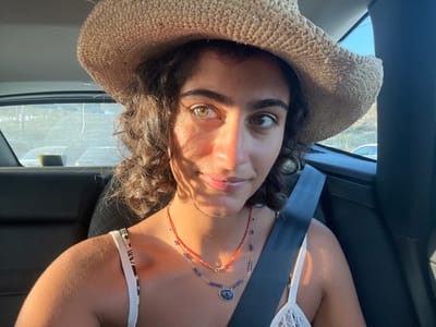 A female individual with dark, curly hair and heterochromatic eyes (one eye appears light-colored, the other darker) is seated in the front passenger seat of a vehicle, described as being in Almancil, Portugal. She is wearing a light-colored straw hat, a white or light-patterned strap top, and two necklaces: one red beaded, and another blue beaded with a central blue pendant. A dark seatbelt is secured across her chest and right shoulder. Direct sunlight illuminates the right side of her face, casting partial shadows on the left. Through the vehicle's rear windows, the exterior environment is visible: to the left, parked vehicles are discernible in an open area, and to the right, a natural, arid-looking hillside with sparse vegetation and a partially visible white vehicle under a clear sky.