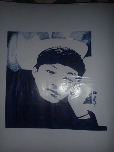 This is a black and white drawing of a person. The person appears to be young, with dark hair and a light complexion. They are wearing a dark garment, possibly a shirt or jacket, and a hat. The drawing is rendered in a sketchy, illustrative style with heavy use of lines and shading. The person's face is the central focus, with their eyes looking directly at the viewer. Their expression is somewhat ambiguous, but could be interpreted as thoughtful or contemplative. The background is plain and undetailed, suggesting the drawing is a portrait. The light source seems to be coming from the right, casting subtle shadows on the person's face and neck. There is a slight sheen on the paper, indicating it might be laminated or covered in protective plastic. No discernible text is present in the image. The drawing is contained within a rectangular frame.