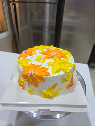 A round cake is positioned on a white cake board, which is then placed on a silver cake stand. The cake is frosted with white buttercream and decorated with an assortment of piped flowers in vibrant shades of orange and yellow. Some of the flowers are adorned with small white and orange edible pearls, and there are scattered yellow sugar pearls across the cake's surface. The background is a blurry indoor setting with a stainless steel appliance, possibly a refrigerator, to the left and a lighter colored wall to the right. The lighting appears to be artificial, casting a soft glow on the cake. There is no indication of the specific city of Dibba Province, Oman.