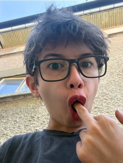 A young person with messy dark hair and black-rimmed glasses is taking a selfie outdoors. They are wearing a dark t-shirt and have their right index finger in their mouth. Their lips are a bright red color, possibly from lipstick or lip gloss. The background is a textured, light-colored wall, and a window with a blue sky reflection is visible to the left. A gutter and a portion of a wooden fence are visible at the top of the image. The scene appears to be in an urban or suburban setting, but no specific location in Peshtera, Bulgaria, can be identified from the visual information alone.