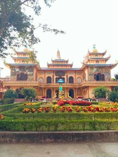 The image depicts a brightly colored Buddhist temple situated in a garden setting in Phú An, Vietnam. The temple features ornate architecture with multiple levels and intricately decorated façades. The building is predominantly painted in hues of light orange and yellow, adorned with detailed artwork and sculptures. The roofs are turquoise. Lush greenery and colorful flowerbeds surround the temple, creating a serene atmosphere. A statue of Buddha sits on a fountain. The sky appears overcast. The ground in the foreground is concrete.