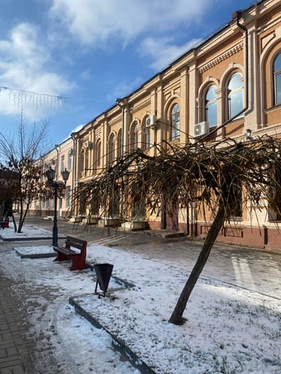 This image captures a quiet winter street scene in Kukui, Russia, under a bright, clear blue sky with scattered white clouds. The ground is partially covered in patchy snow, mixed with visible pavement and some grassy areas.

A prominent feature is a large, multi-story building constructed of light brown or beige brick, situated on the left side of the frame. The building exhibits a classical architectural style, characterized by numerous arched windows with decorative molding above them, and detailed cornices and brickwork along its facade. Several white air conditioning units are mounted beneath some of the windows.

In the foreground and center, a large, bare-branched tree with a wide, sprawling canopy stands beside the building, its intricate network of branches silhouetted. Further down the street, another smaller, bare tree is visible. The street area itself consists of paved sections and areas with melting snow. A red-sided wooden park bench and a black metal trash can are positioned on the snowy ground. Several dark, ornate lampposts are spaced along the street. A string of decorative lights hangs overhead, stretching across the scene against the sky. The bright sunlight casts distinct shadows on the snow and ground, indicating it is daytime.

No people or activities are visible in the image, contributing to a peaceful and still atmosphere. There is no discernible text visible on any objects within the frame.