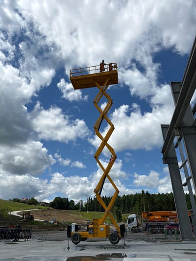 The image depicts a construction site under a cloudy sky. A tall, yellow scissor lift dominates the center of the frame, its platform elevated high in the air. Two construction workers, wearing high-visibility vests, are standing on the platform of the scissor lift. One worker is operating the machinery, while the other appears to be observing or assisting. To the right of the scissor lift, a partially constructed steel building frame is visible, suggesting ongoing construction work. In the background, a wooded hillside with a winding road and scattered houses can be seen, indicative of a rural or semi-rural setting outside of a densely populated urban area. A truck with an extended boom is parked behind a barrier, and other construction equipment, including excavators and barriers, are present on the ground. The presence of construction equipment, workers, and unfinished structures clearly indicates an active construction site.