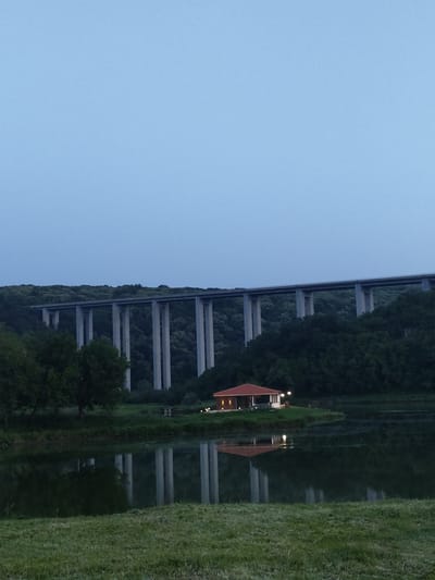 This image, captured in Pisanets, Bulgaria, depicts a landscape scene at twilight. A multi-span concrete viaduct, light grey in color, traverses a densely forested hillside in the background. A calm, dark body of water occupies the mid-ground, reflecting the viaduct's piers and a small structure. This single-story building, situated on a small landmass extending into the water, features a red-orange tiled roof and light-colored walls. Multiple exterior lights on the building are illuminated, casting warm yellow glows and reflecting on the water's surface. The foreground consists of light green grass, with additional trees visible near the water's edge. The sky overhead is a clear, light blue. No individuals are visible.