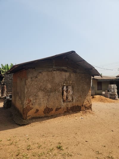 The image displays a weathered, single-story building under a clear, bright sky, set on dry, light brown ground. The structure, characteristic of a rural or semi-urban environment like Umuoye, Nigeria, shows significant signs of age and disrepair. Its walls, likely made of concrete or plastered earth, have large sections where the outer rendering has peeled away, exposing a darker, reddish-brown material underneath. A small, rectangular opening on the side wall is covered by a worn wooden panel or shutter, serving as a window. The roof consists of corrugated metal sheets, appearing rusty.
The surrounding ground is predominantly sandy dirt, with sparse patches of green vegetation scattered, particularly visible towards the right side. In the background, to the right of the building, a stack of concrete blocks or similar construction materials can be observed. Hints of large green foliage, possibly from palm trees, are visible in the far left background. The strong, direct sunlight indicates a clear day, casting defined shadows. No people or visible text are present in the scene.
