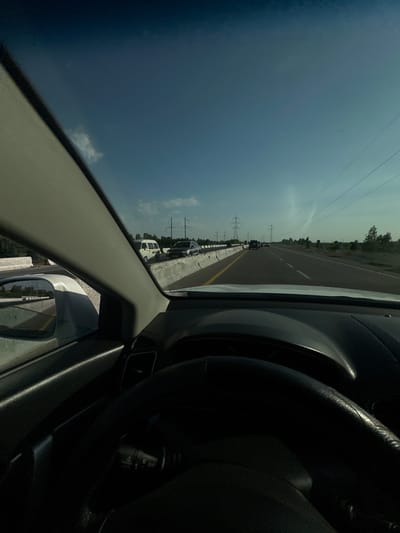 The image is taken from the interior of a car, looking forward through the windshield and out the passenger side window. The perspective suggests the car is driving on a highway with multiple lanes, currently experiencing moderate traffic. The sky is a clear, pale blue with some wispy clouds. Power lines and transmission towers are visible along the side of the road, indicating a developed area. Trees and vegetation line the roadside in the distance.
In the foreground, the steering wheel and dashboard of the car are partially visible, along with the driver's side windshield pillar. To the left, the side mirror reflects a portion of the road and the surrounding landscape, showing a white vehicle in the adjacent lane. On the highway ahead, several vehicles are present, including a white van and a dark-colored sedan. The road surface appears to be asphalt, with white and yellow lane markings. The overall impression is a travel scene on a sunny day in a semi-urban or rural environment.
The location is identified as a highway in the city of Istiklol, Uzbekistan.