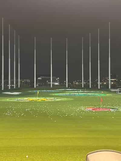 The image depicts a night-time view of a Topgolf facility, specifically the driving range area, in Schaumburg, Illinois, United States of America. Tall, brightly lit poles, characteristic of Topgolf's setup, dominate the background. In the mid-ground, the expansive green turf of the driving range is visible, dotted with numerous white golf balls. Several target areas are illuminated with colored lights, and flags mark some of these targets, including a yellow flag and a red flag. In the further distance, buildings and city lights are visible, suggesting an urban or suburban setting. The foreground shows a portion of a golf cart or similar seating, implying a viewer's perspective from a bay. The overall scene is that of a popular recreational facility operating after dark.
