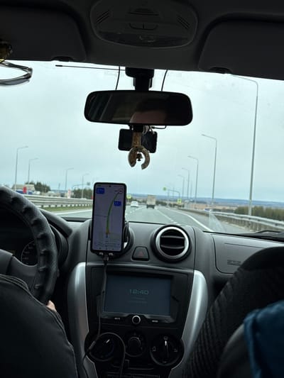 The image provides a driver's perspective from inside a car traveling on a multi-lane highway or wide road, likely in Покровское, Russia. The sky outside appears overcast and bright, indicating daytime.
The primary subjects include the car's interior and the road ahead. A driver's arm and leg are visible on the right, with their hand resting on a black, perforated steering wheel. A smartphone, mounted centrally on the dashboard, displays a navigation application showing a route. The navigation indicates "178 км" (178 km), an estimated arrival time of "14:40", and "2 ч 1 мин" (2 hours 1 minute) remaining. The phone's screen also shows a current speed of "50" (likely km/h) and a speed limit of "11".
On the car's built-in display, the time "12:40", the day "пятница" (Friday), and the date "01.05 2026" are visible. Below this, buttons for "MUTE" and "MODE" are present, along with climate control knobs. An "SRS AIRBAG" label is on the passenger side of the dashboard. A rearview mirror hangs in the center, reflecting a blurred image, possibly of the driver. A small, metallic charm, possibly a horseshoe shape, displaying "1487" or "14871" is attached near the rearview mirror.
The scene depicts a typical long-distance journey, with the driver using navigation for guidance. The presence of streetlights or gantries lining the road suggests a developed infrastructure. The time difference between the car's clock (12:40) and the navigation's estimated arrival time (14:40) is notable, possibly due to time zone settings or an incorrectly set car clock given the future date.