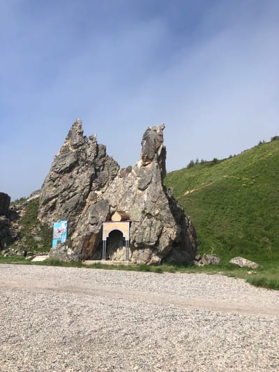 An outdoor, eye-level, wide shot captures a religious shrine built into a natural rock formation in Ola, Russia. The shrine, an arched structure with a golden roof, is situated on the face of a large, jagged rock. To the left of the shrine, a blue and white mural is attached to the rock. A path of gravel extends from the foreground into the middle ground. The background features a lush green hillside with scattered trees and a clear blue sky. A bird is perched on the highest point of the rock formation. The scene is lit by daylight, and the weather appears clear.