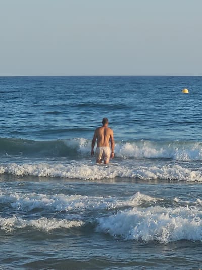 A person stands in the ocean facing away from the camera, with waves breaking around them. The person is wearing striped swim trunks. A yellow buoy is visible in the upper left corner of the frame. The scene appears to be a beach on a sunny day. The water is a deep blue, and the waves are white and foamy. The sky is a light blue. The location is Castelldefels, Spain.