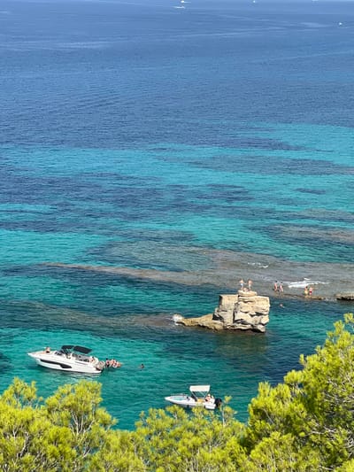 This image depicts a coastal area near Es Caló de Sant Antoni, Llucmajor, Spain, viewed from an elevated position. The foreground is occupied by green foliage from a tree. The majority of the frame consists of clear ocean water, displaying transitions from light turquoise in shallower areas to deep blue in the distance.

In the mid-ground, a substantial, light-colored rock stack rises from the water. Two individuals stand on the flat top of this stack. To the right of the stack, a shallow rock platform is visible, where approximately four individuals are standing or wading in the water. Extensive submerged rock formations and reefs are discernible beneath the water's surface around these features.

Two white motorboats are present in the water in the lower-mid ground. A larger motorboat with a black bimini top is positioned on the left, with several individuals in the water around its stern, including one on a circular inflatable raft. A smaller motorboat with a light-colored bimini top is located to the right of the larger vessel. Individuals are observed swimming or engaged in recreational activities in the water surrounding both boats and the rock formations. In the far background, across the deeper blue expanse of the sea, several indistinct white vessels are visible on the horizon.