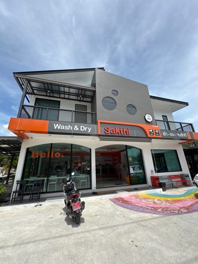 A sideways shot depicts the exterior of "Sakthi Wash & Dry", a laundromat located in Moo 4, Thailand. The modern, two-story building features a gray facade with circular windows and an orange vertical sign with the business name written in English and Thai, along with "Wash & Dry". Large glass windows reveal orange washing machines inside. A motorcycle with a Thai license plate is parked in front. A faded rainbow design is painted on the ground adjacent to the building. The sky above is blue with scattered clouds.
